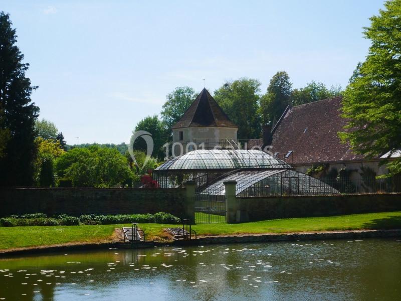 Vue d'une serre en verre et d'un bâtiment ancien entourés de verdure, près d'un étang calme.