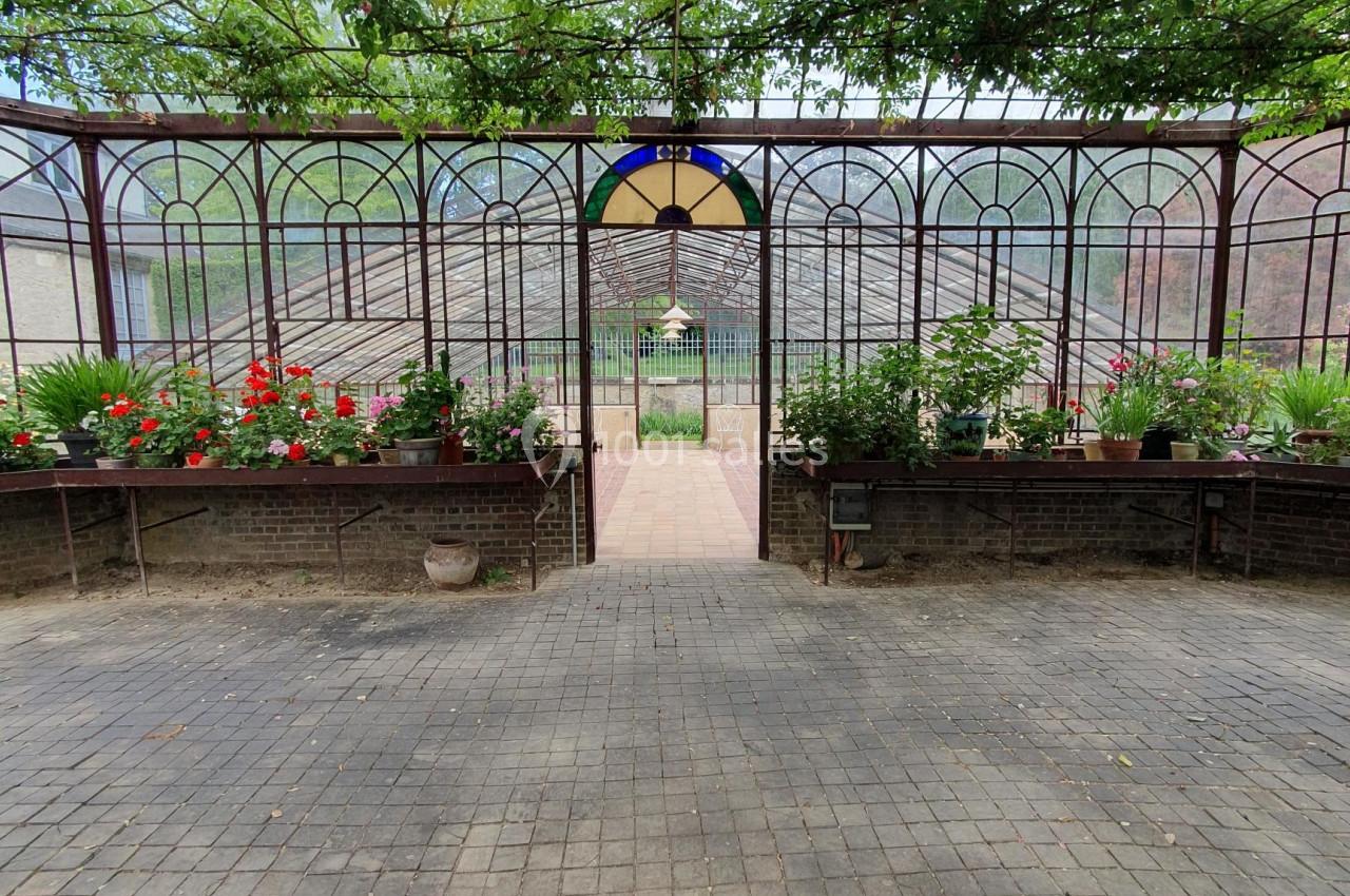 Serre avec structure métallique, plantes fleuries et allée pavée menant à une porte ouverte sur un jardin.