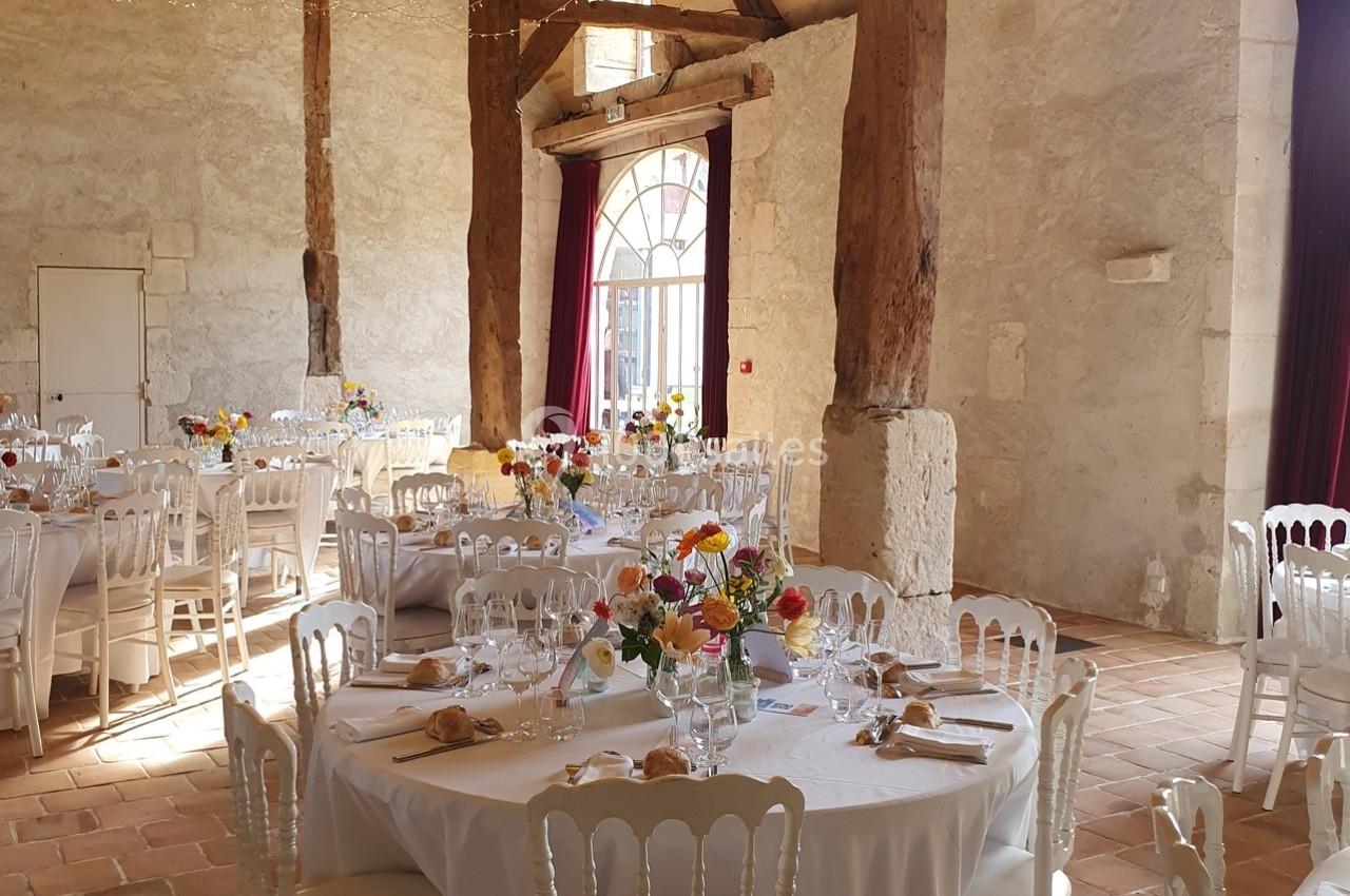 Salle de réception rustique avec tables rondes dressées, décorées de bouquets colorés et chaises blanches.