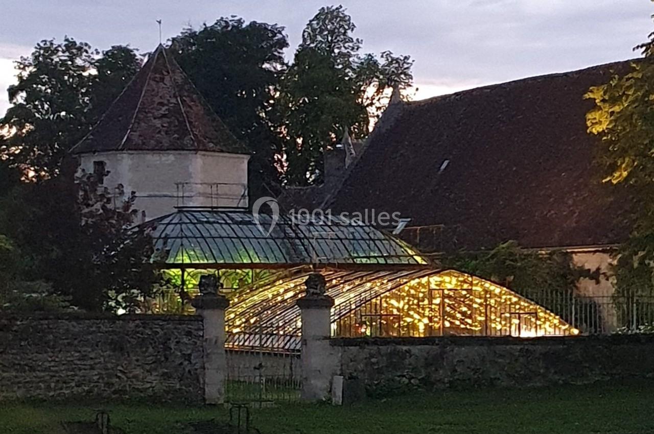 Serre illuminée au crépuscule, entourée de murs en pierre et d'arbres, avec une tour en arrière-plan.