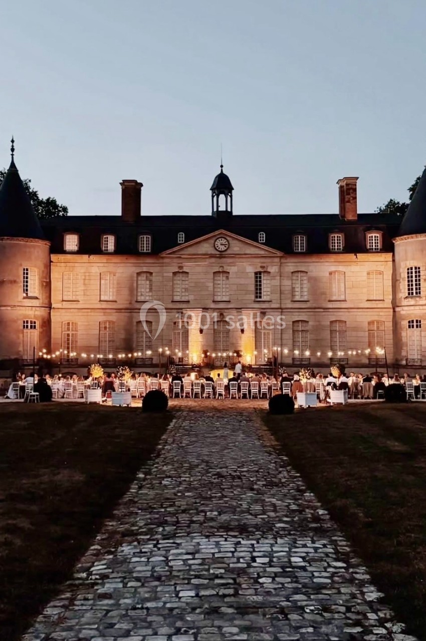 Château éclairé au crépuscule avec des tables dressées et des invités réunis dans la cour pour un événement en plein air.
