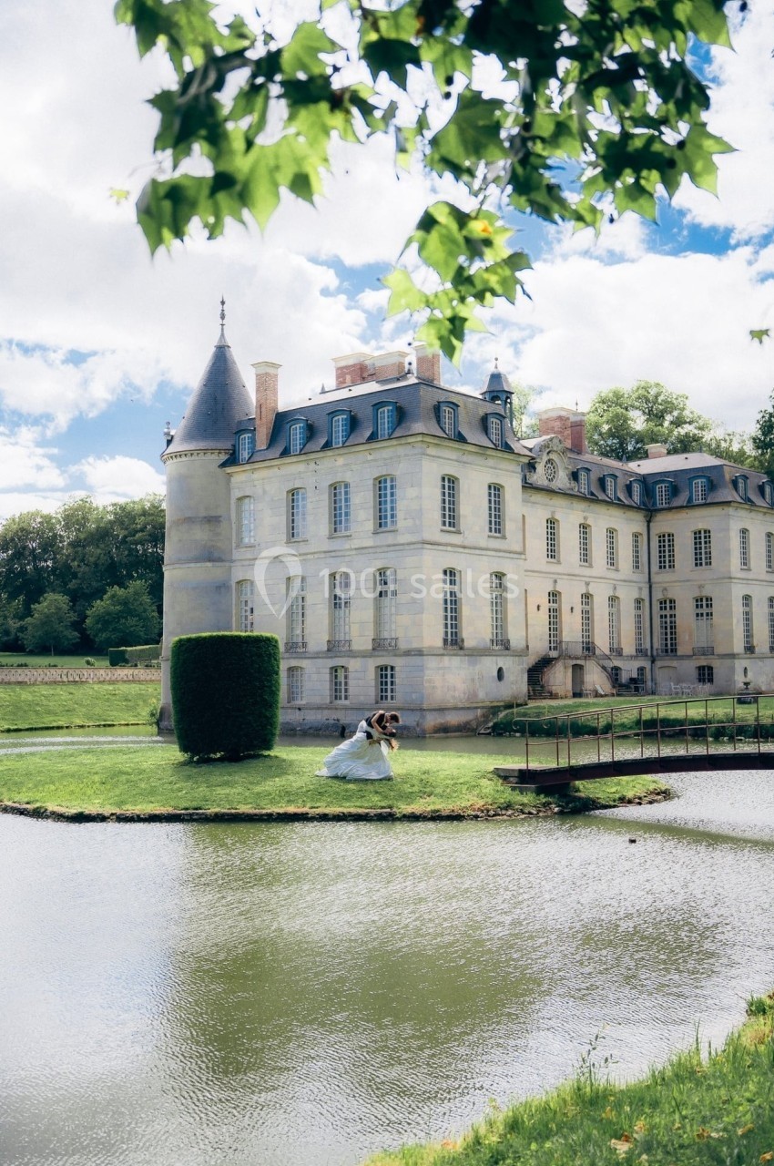 Château historique entouré d'un parc verdoyant avec un étang au premier plan, sous un ciel partiellement nuageux.