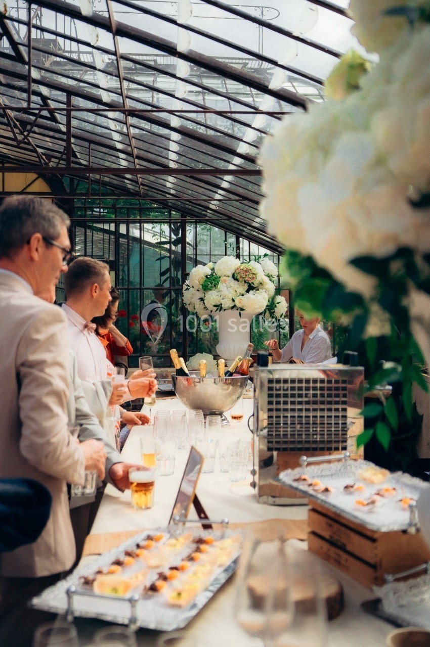 Des invités discutent autour d'une table garnie de boissons et d'amuse-bouches dans une serre lumineuse.