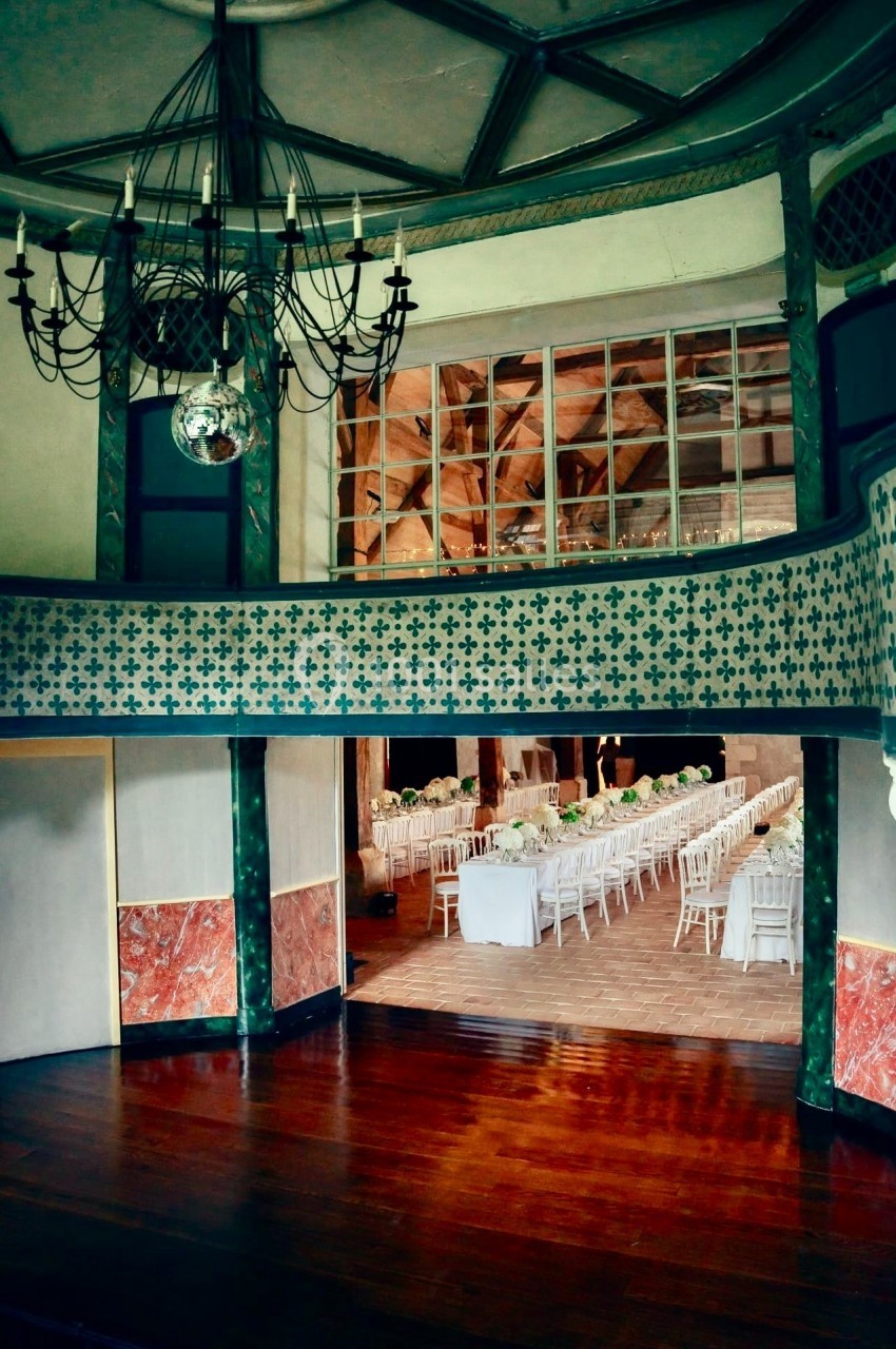 Salle de réception avec parquet, lustre, balcon décoratif et tables dressées pour un événement.