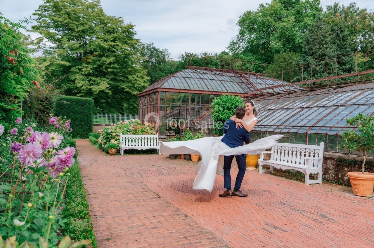 Un couple en tenue de mariage danse dans une allée pavée près de serres entourées de verdure et de fleurs.