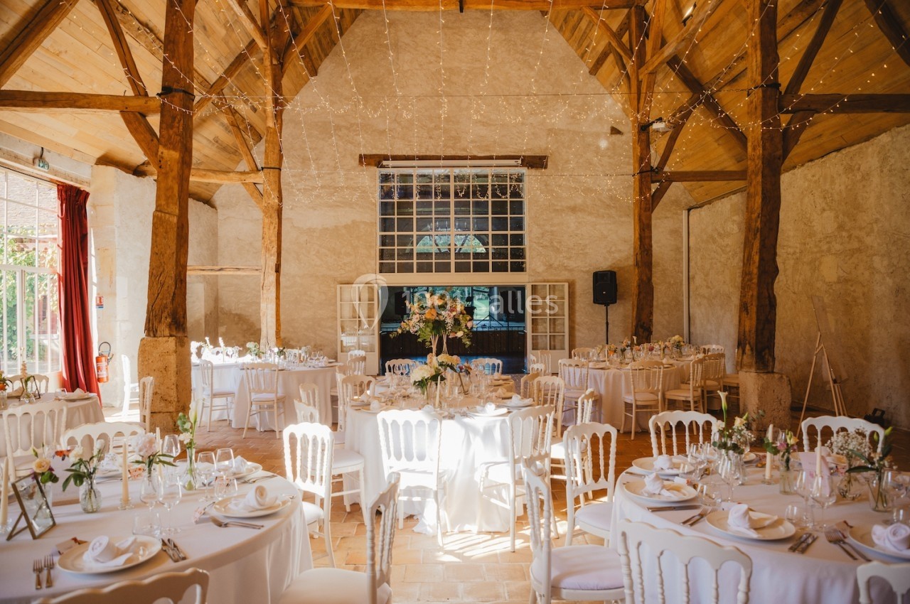 Salle de réception lumineuse avec tables rondes décorées pour un mariage, sous un plafond en bois avec guirlandes lumineuses.