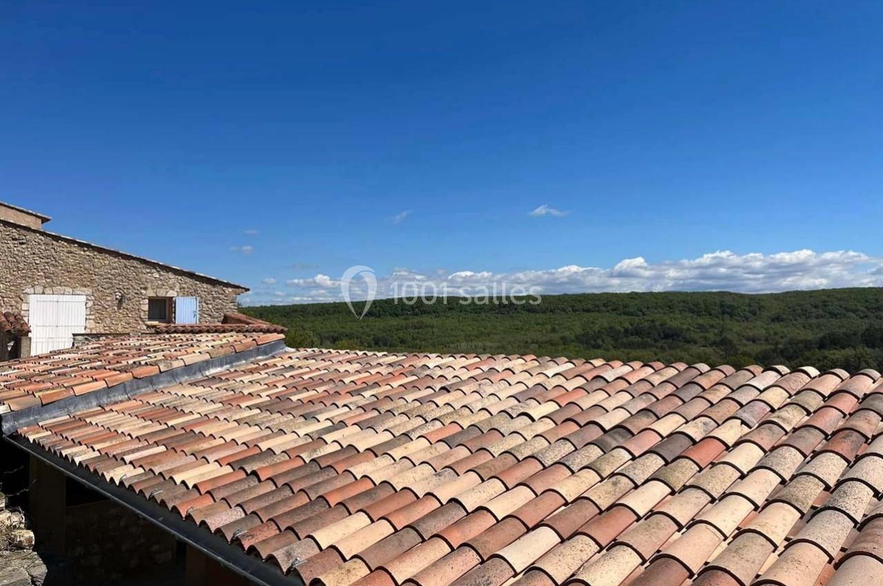 Toits en tuiles rouges d'une maison en pierre, avec une vue dégagée sur une forêt et un ciel bleu.