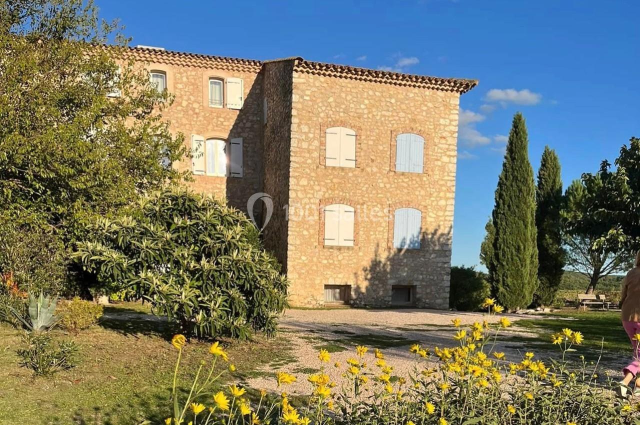 Bâtisse en pierre avec volets bleus entourée de végétation, sous un ciel bleu dégagé.