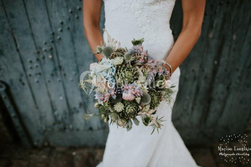 Une mariée en robe blanche tient un bouquet de fleurs pastel devant une porte en bois bleu vieilli.