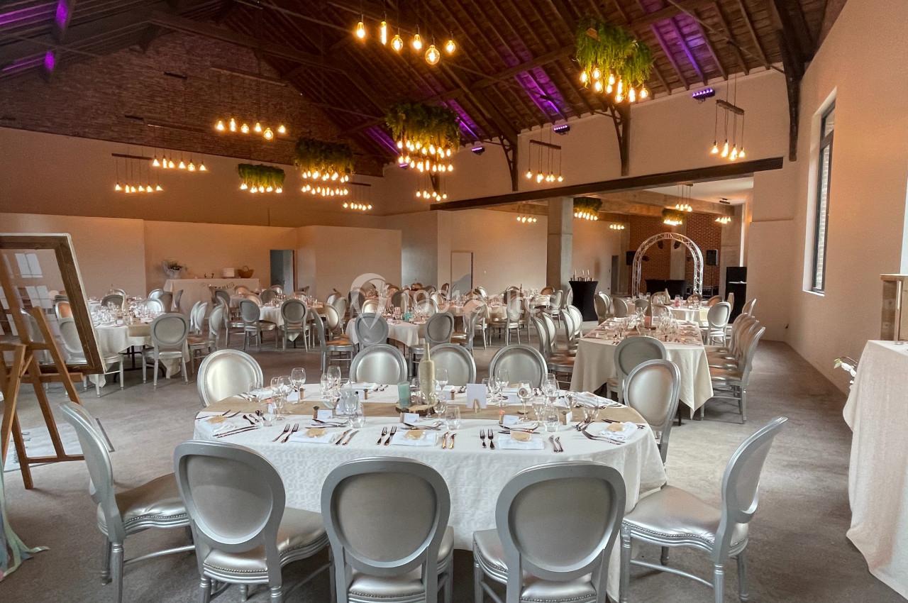 Salle de réception décorée avec des tables rondes dressées, des chaises argentées et des luminaires suspendus.