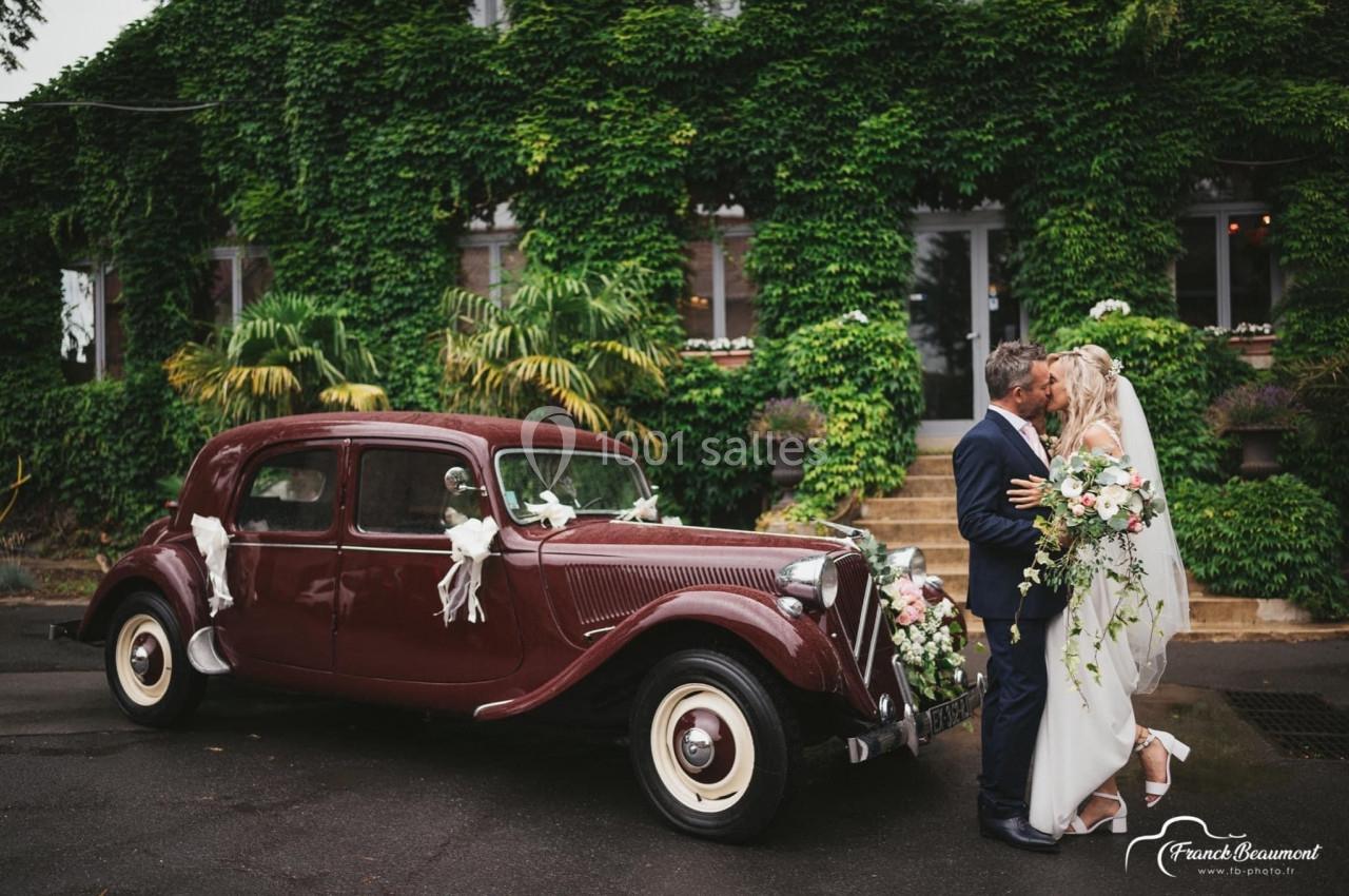 Un couple de mariés s'embrasse devant une voiture ancienne décorée, avec un bâtiment couvert de verdure en arrière-plan.