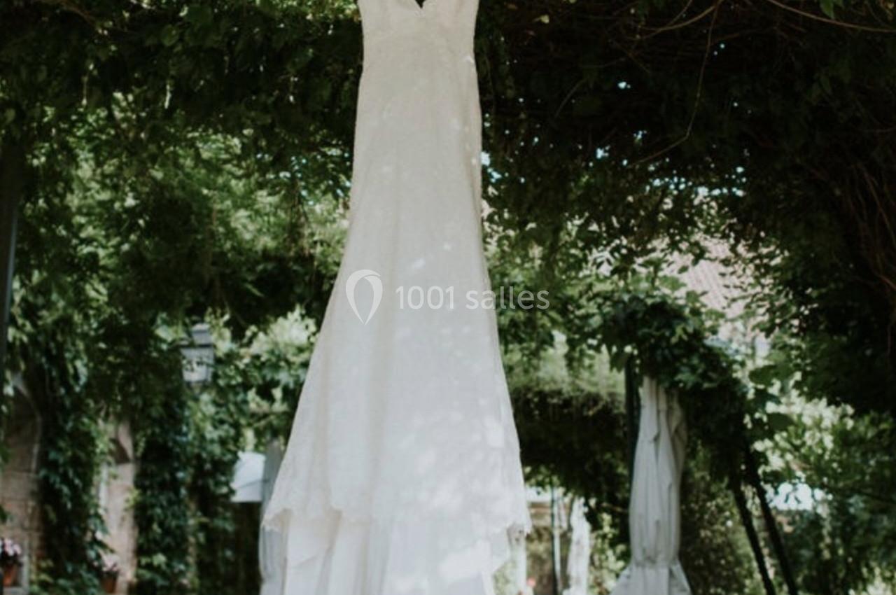 Robe de mariée blanche suspendue à une branche sous une pergola verdoyante avec des tables et chaises en fer forgé.