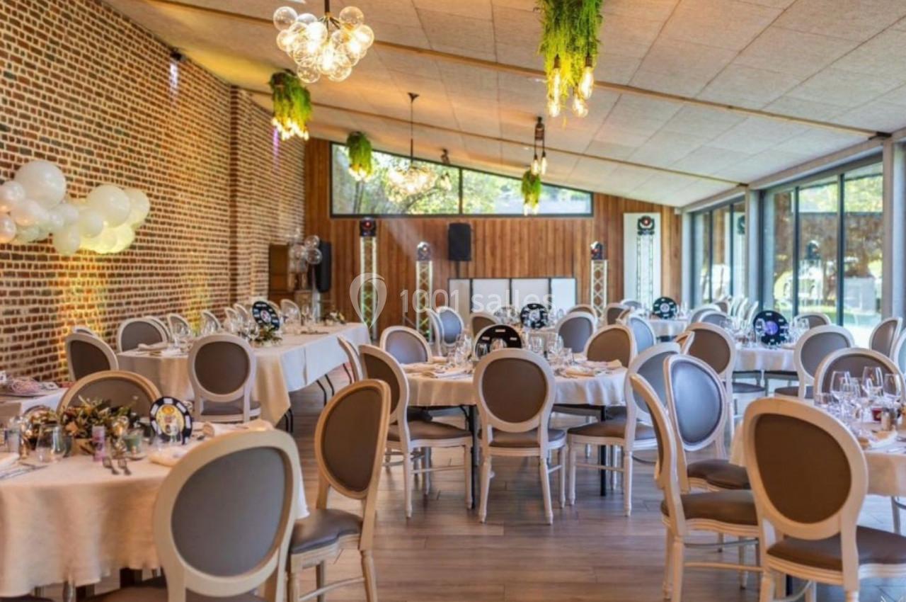 Salle de réception lumineuse avec tables rondes dressées, murs en briques, grandes baies vitrées et décorations suspendues.
