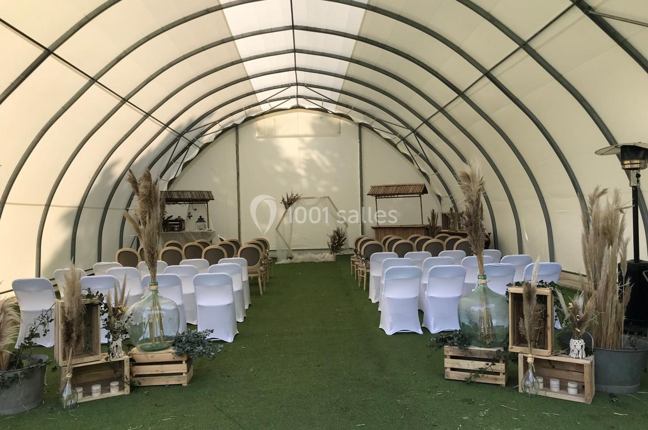 Salle de cérémonie décorée avec des chaises blanches, des éléments en bois, des plantes et des jarres en verre.