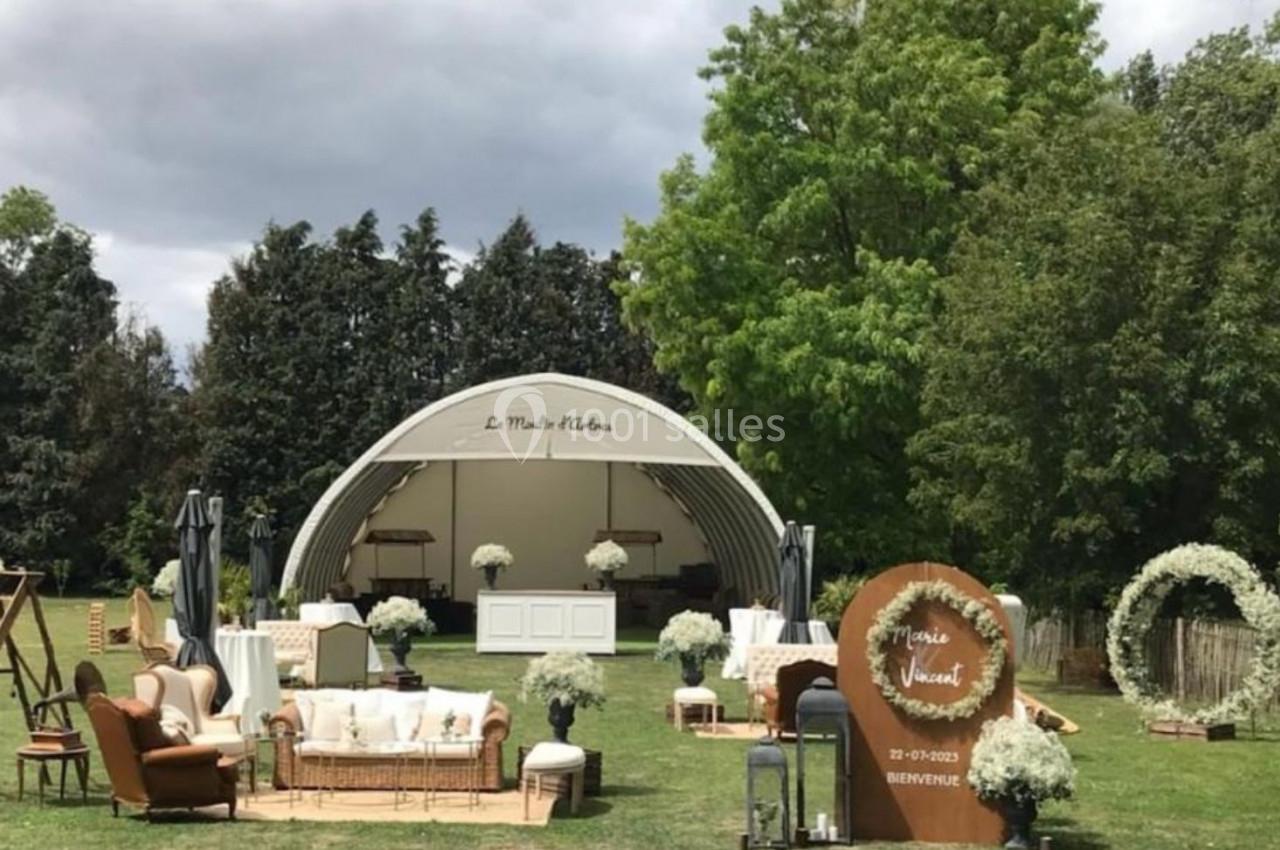 Espace extérieur aménagé pour un événement, avec une scène couverte, des fauteuils, des décorations florales et des arbres…