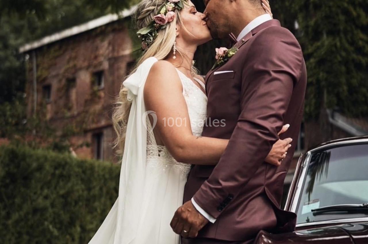 Un couple en tenue de mariage s'embrasse devant une voiture ancienne, avec un bâtiment en briques en arrière-plan.