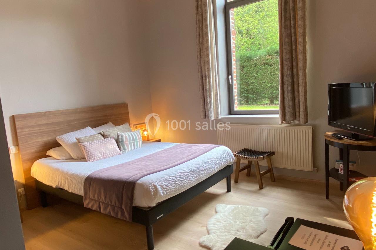 Chambre lumineuse avec lit double, bureau, lampe suspendue et grande fenêtre donnant sur un jardin verdoyant.