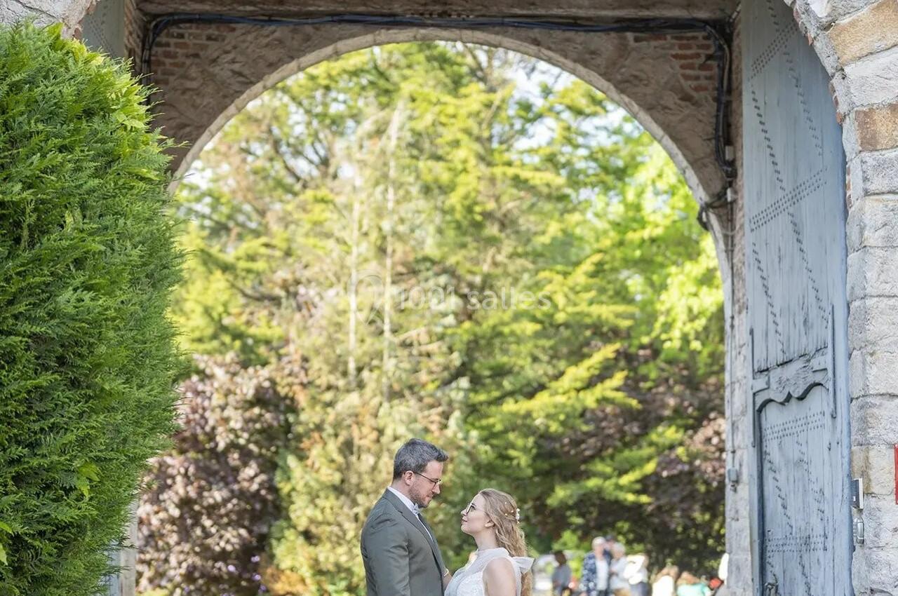 Un couple de mariés se tient sous une arche en pierre dans un jardin verdoyant par une journée ensoleillée.