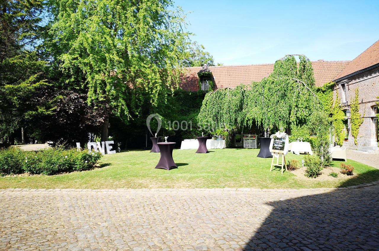 Cour extérieure avec pelouse, tables hautes noires, arbres et bâtiments en briques sous un ciel bleu.