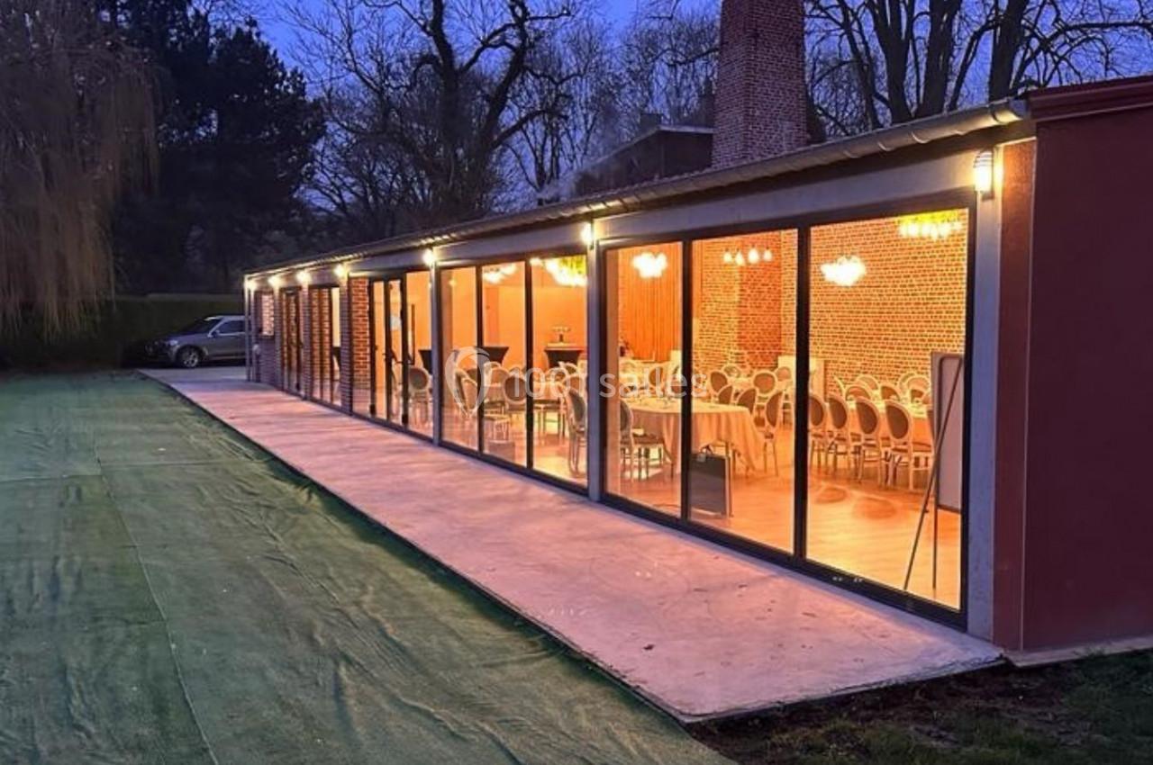 Salle vitrée éclairée en soirée, avec des tables et chaises visibles à l'intérieur, entourée d'un espace extérieur herbeux.