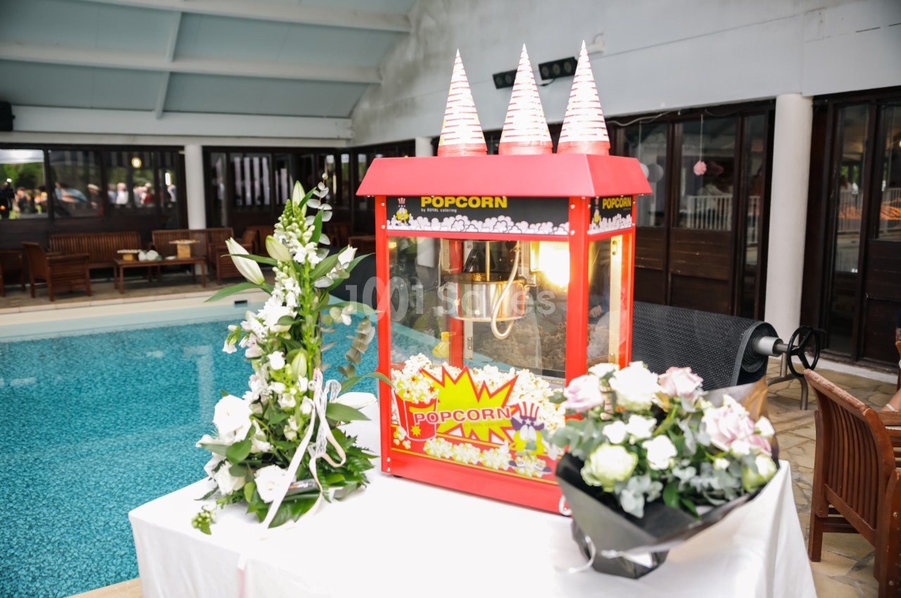 Machine à pop-corn rouge décorée, entourée de fleurs blanches et roses, placée près d'une piscine intérieure.