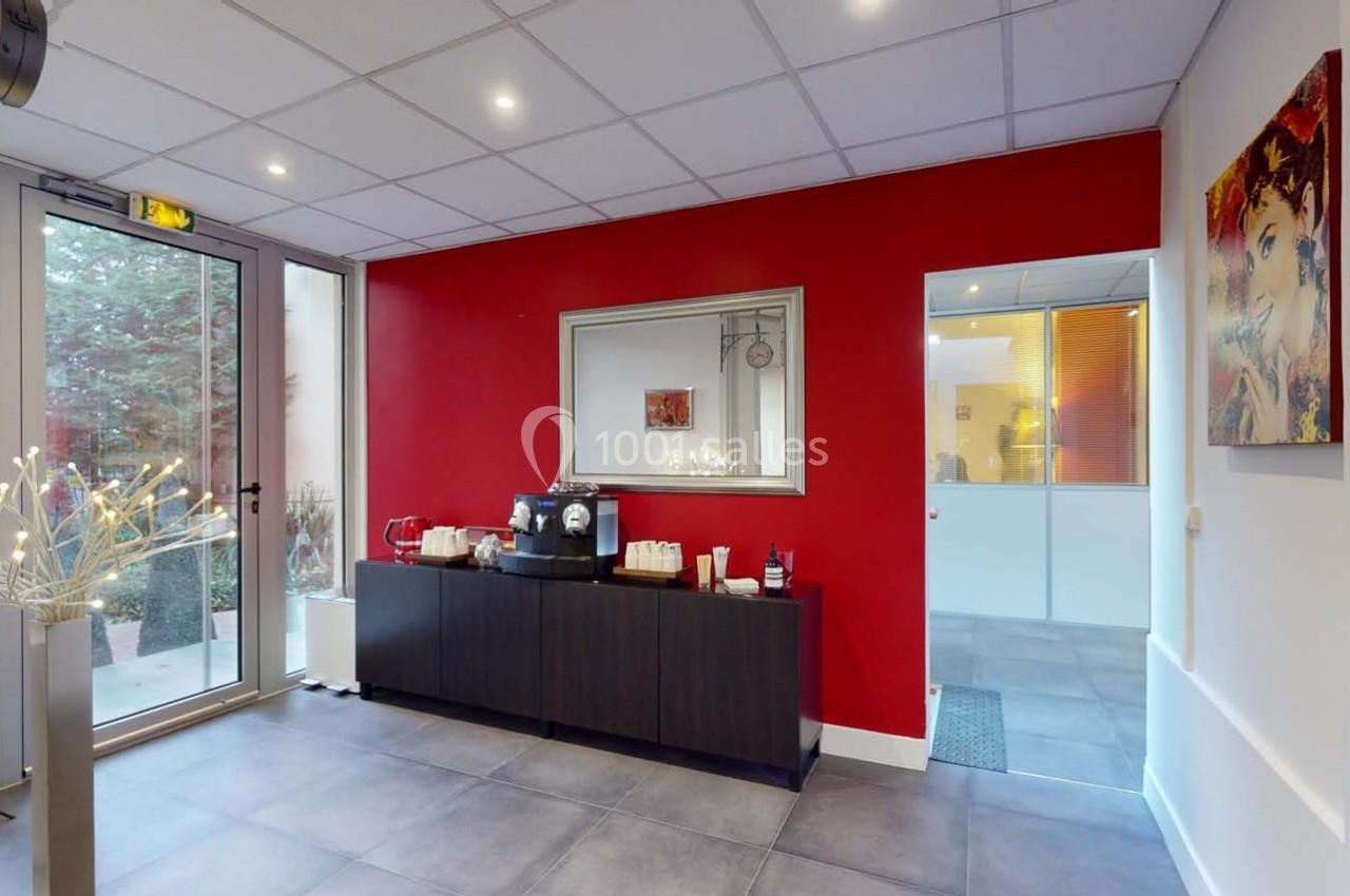 Espace d'accueil lumineux avec mur rouge, machine à café sur meuble, et porte vitrée donnant sur l'extérieur.
