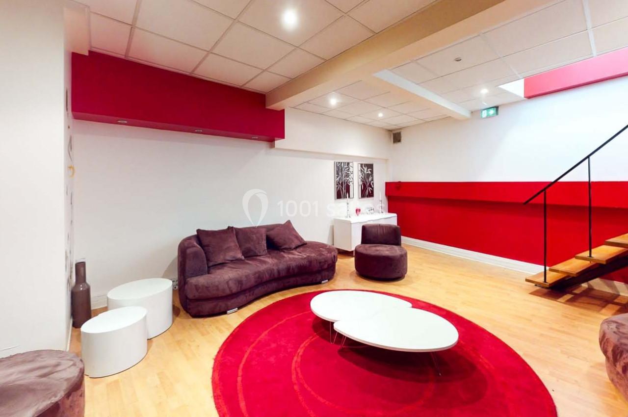 Salon moderne avec canapé violet, tables basses blanches, tapis rouge rond et escalier en bois dans un espace lumineux.