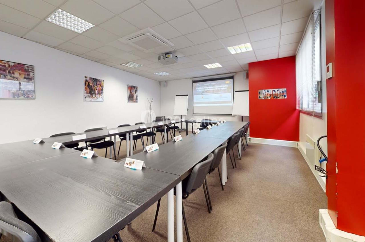 Salle de réunion équipée de tables noires disposées en U, chaises, vidéoprojecteur et tableau blanc.