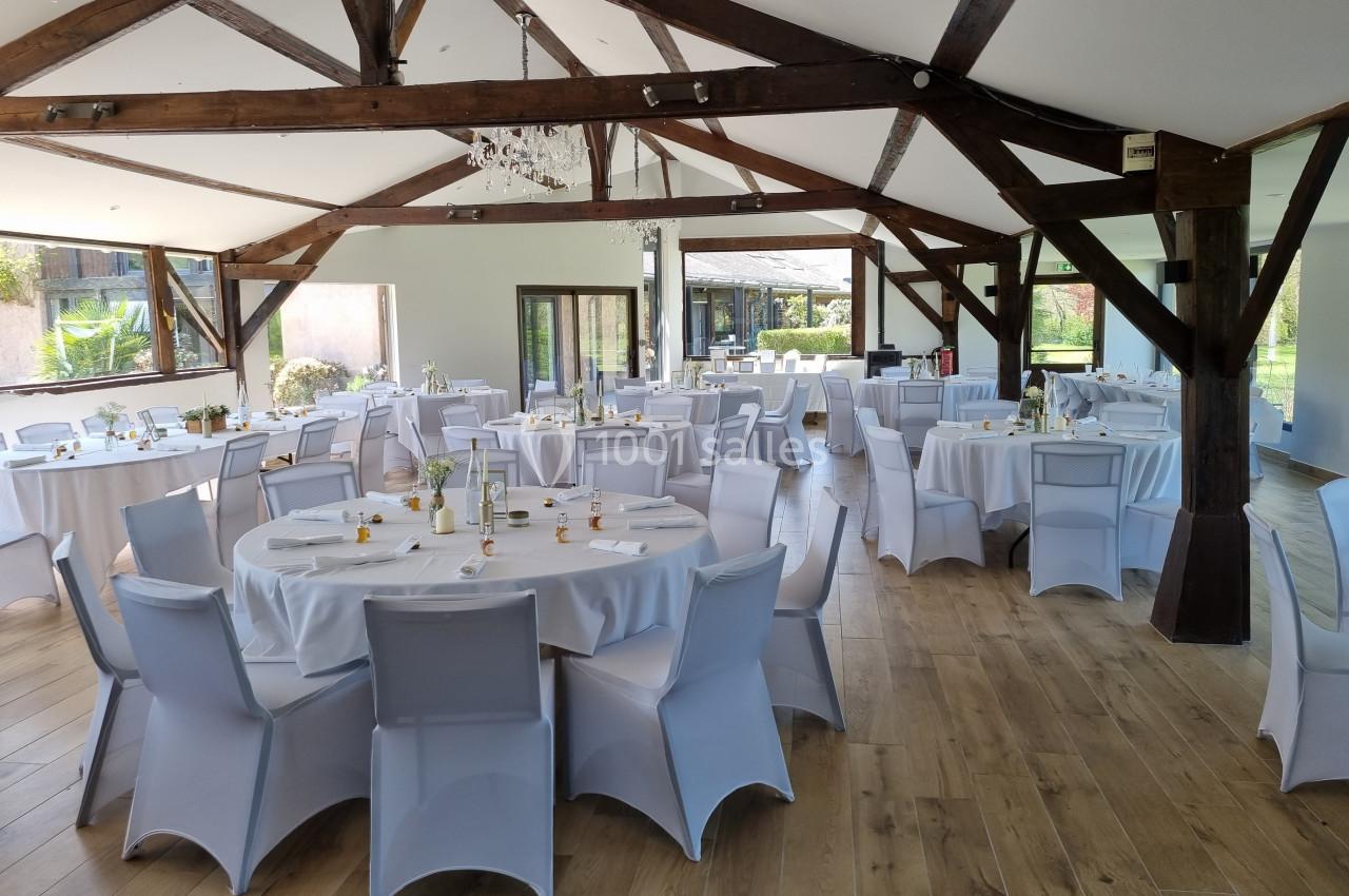 Salle de réception lumineuse avec tables rondes dressées, chaises blanches et poutres apparentes en bois.