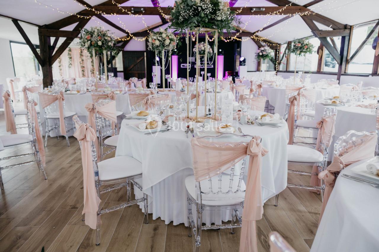 Salle de réception décorée pour un mariage avec des tables rondes, nappes blanches et chaises ornées de rubans roses.