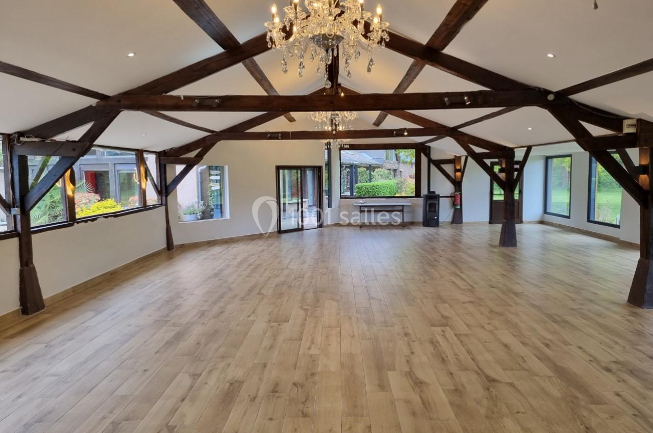 Salle spacieuse avec parquet en bois clair, poutres apparentes et lustre, entourée de grandes fenêtres donnant sur un jardin.