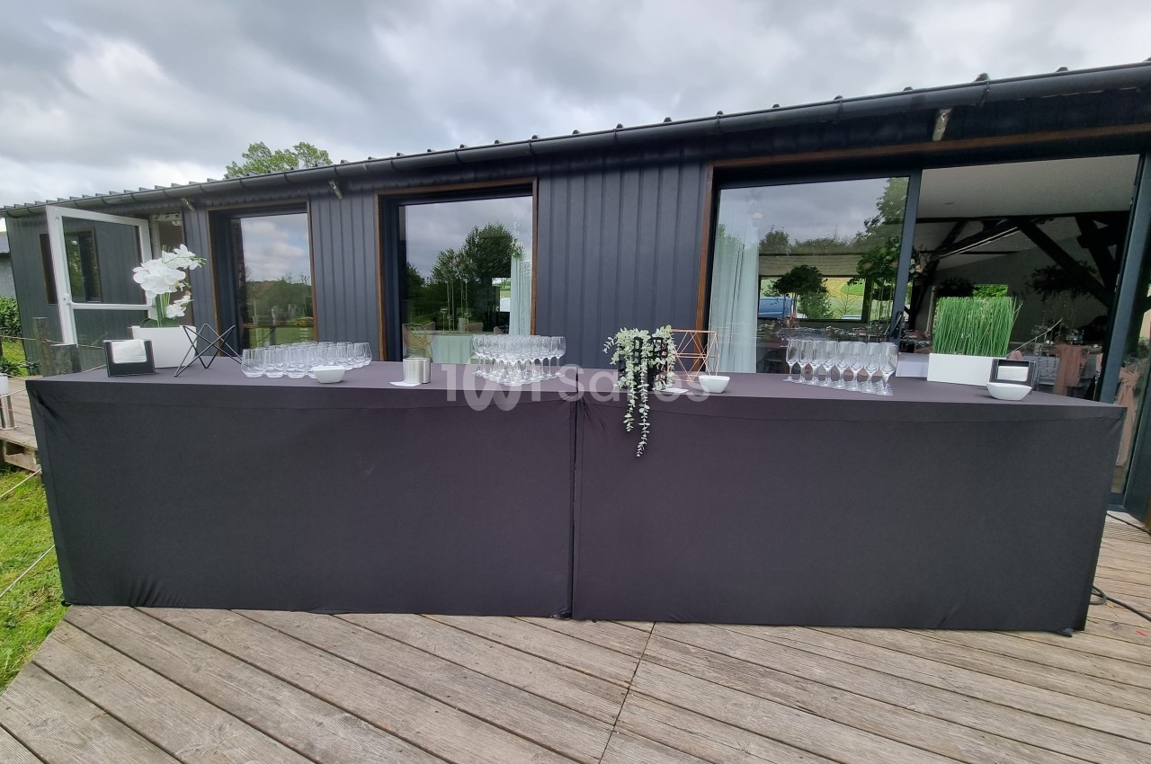 Table de service extérieure avec nappes noires, verres alignés et décoration florale, devant un bâtiment moderne.