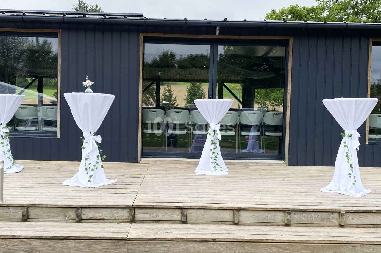 Tables hautes décorées de nappes blanches et de feuillages, disposées sur une terrasse en bois devant un bâtiment vitré.