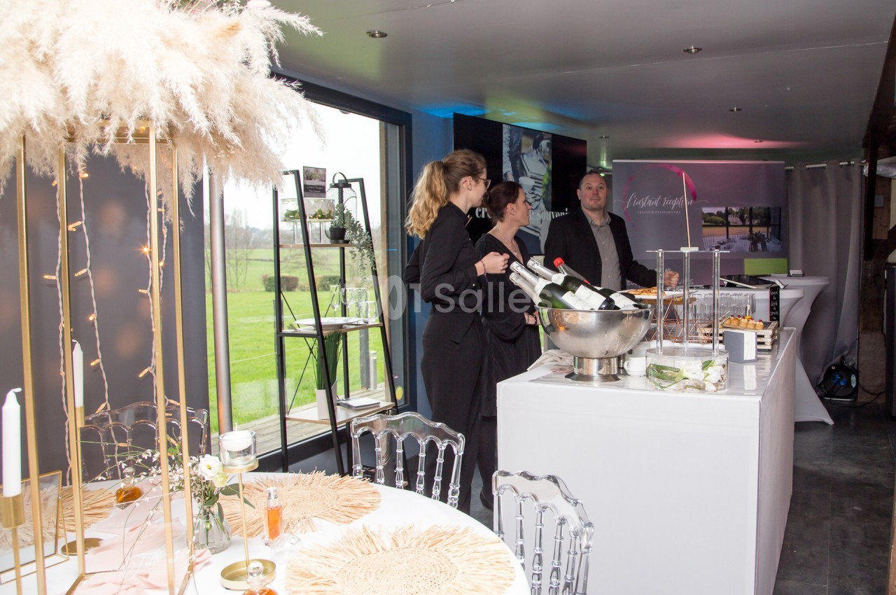 Trois personnes discutent près d'un bar avec des bouteilles de champagne, dans une salle décorée pour un événement.