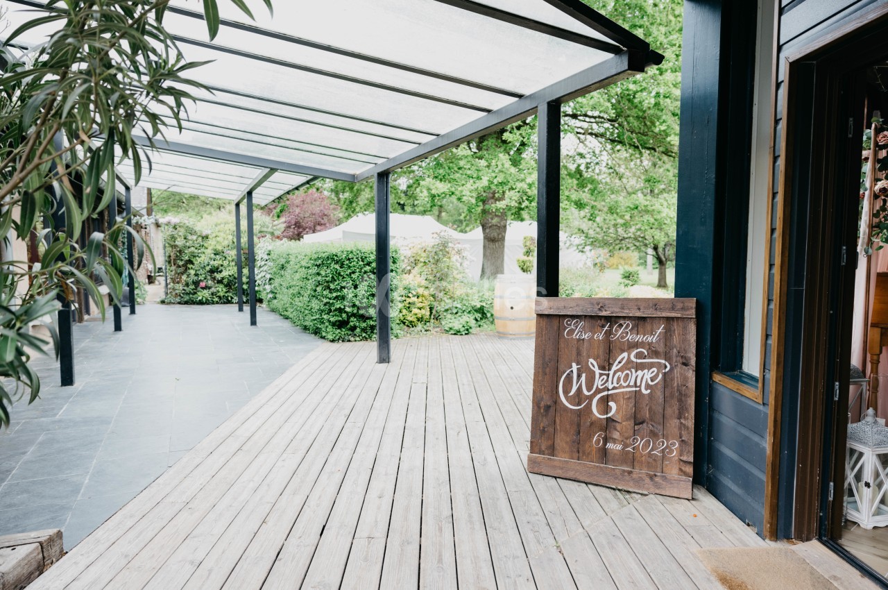 Terrasse en bois couverte, entourée de verdure, avec un panneau de bienvenue indiquant une date.