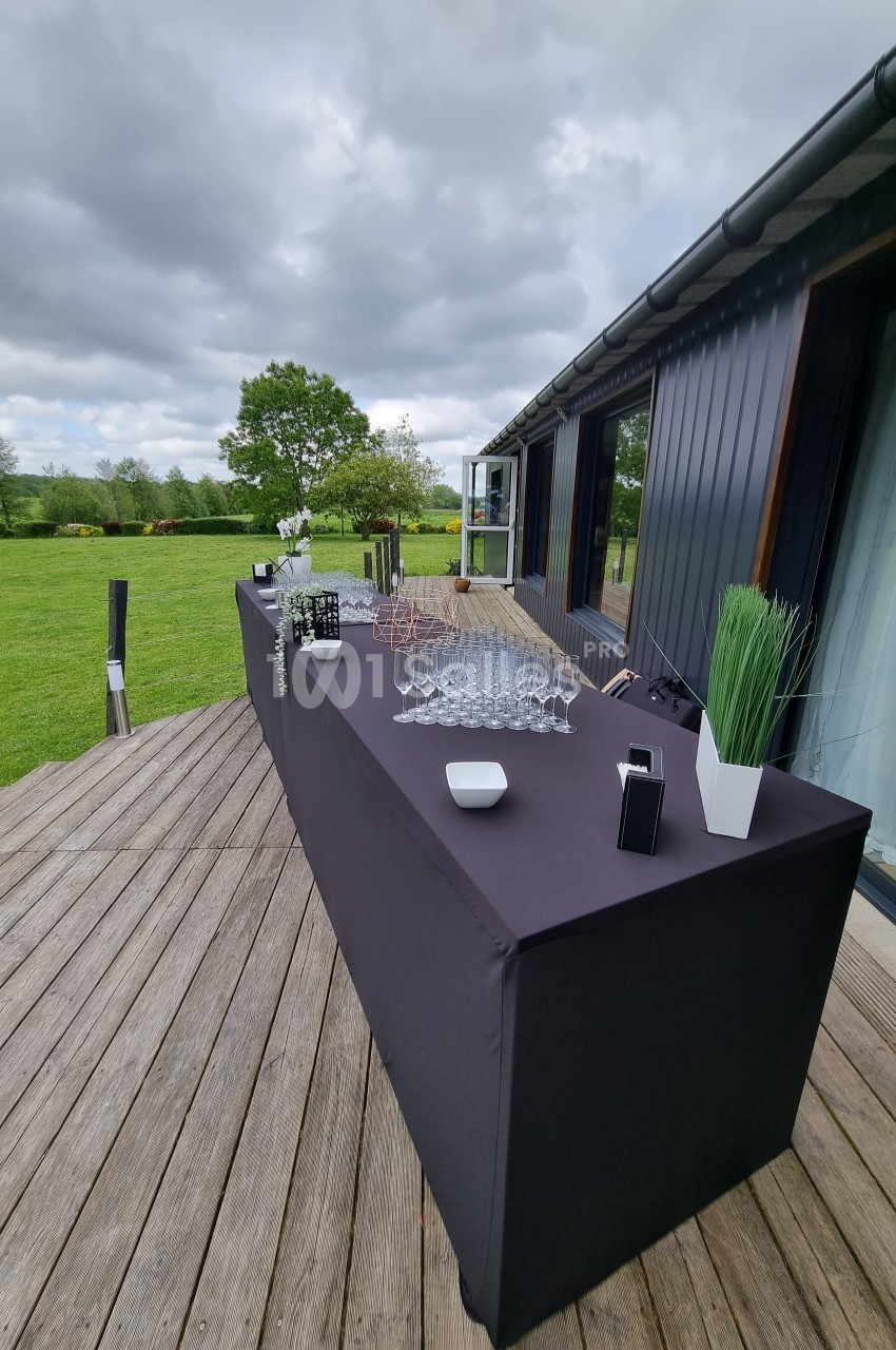 Table dressée en extérieur sur une terrasse en bois, avec verres, bouteilles et décorations, sous un ciel nuageux.