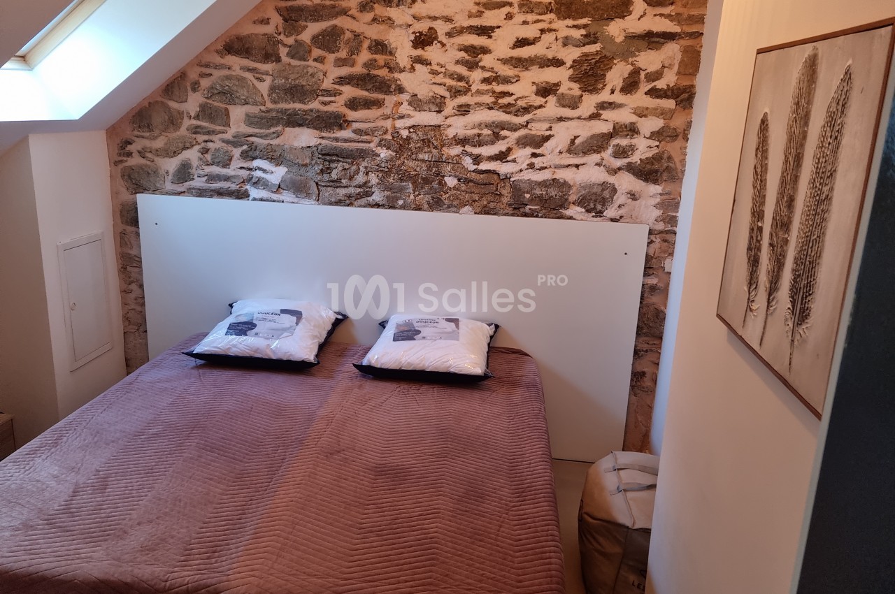 Lit double avec couverture marron dans une chambre mansardée, mur en pierres apparentes et tableau décoratif.