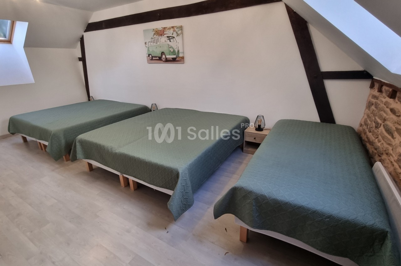 Trois lits simples avec couvre-lits verts dans une chambre mansardée aux murs blancs et poutres apparentes.
