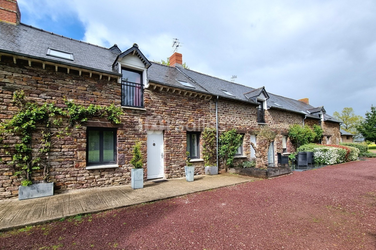 Façade en pierre d'une maison traditionnelle avec volets noirs, entourée de végétation et d'une allée en gravier rouge.