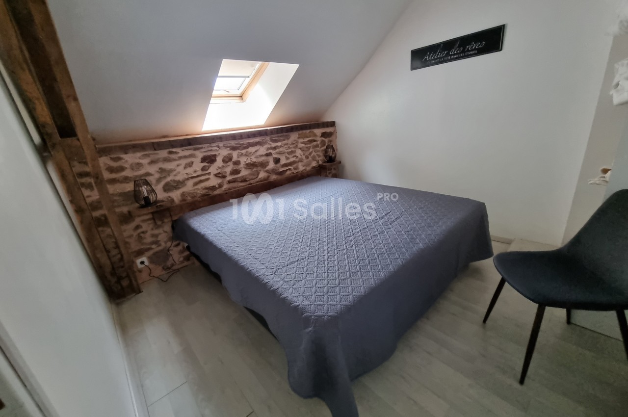 Chambre mansardée avec lit double, mur en pierre apparente, fenêtre de toit et chaise grise sur sol clair.
