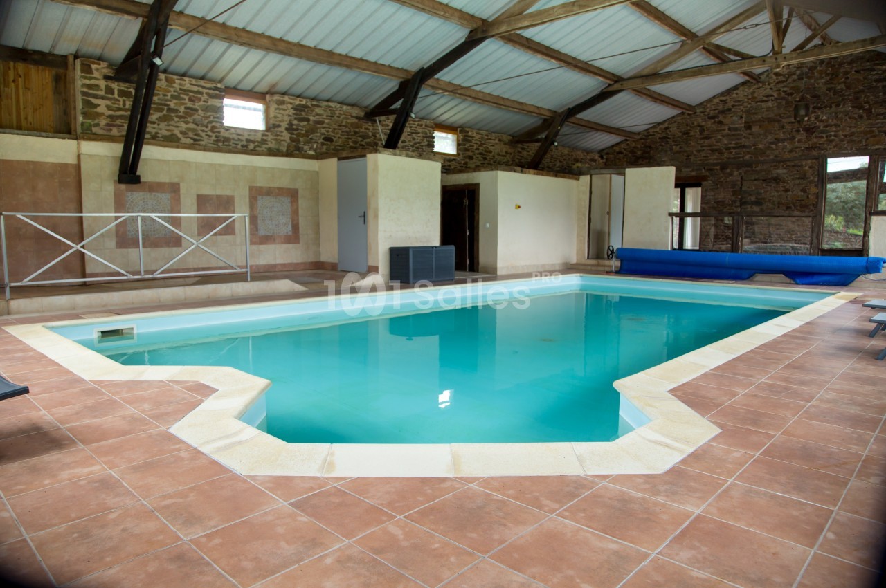Piscine intérieure avec carrelage beige, entourée de murs en pierre et d'une charpente en bois sous un toit en tôle.
