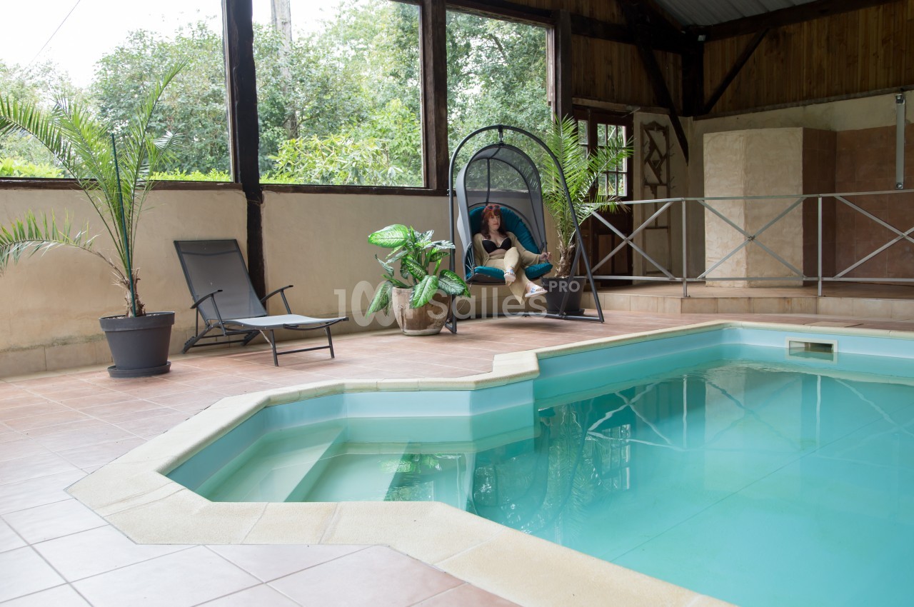 Piscine intérieure avec carrelage beige, transat, plantes en pot et une personne assise dans un fauteuil suspendu.