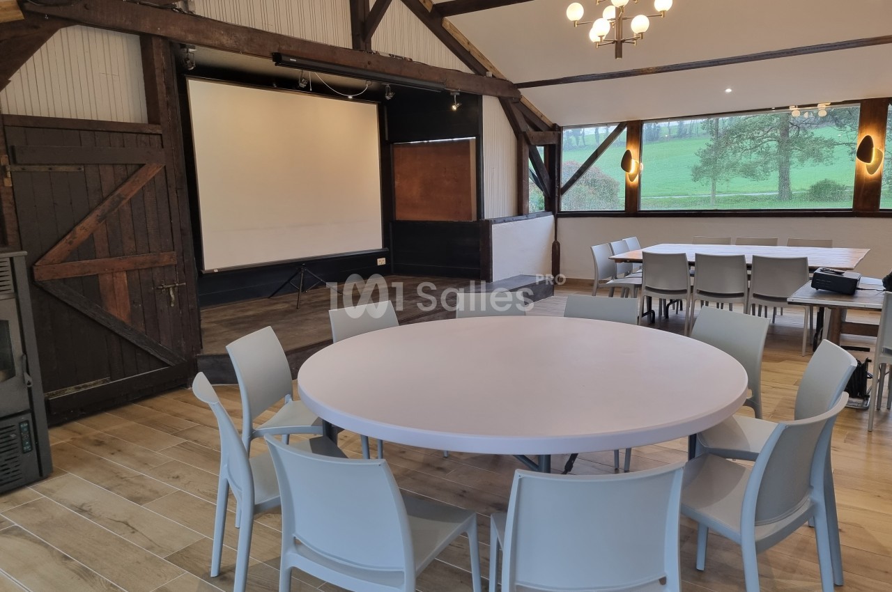 Salle de réunion avec tables rondes, chaises, écran de projection et vue sur un paysage extérieur verdoyant.