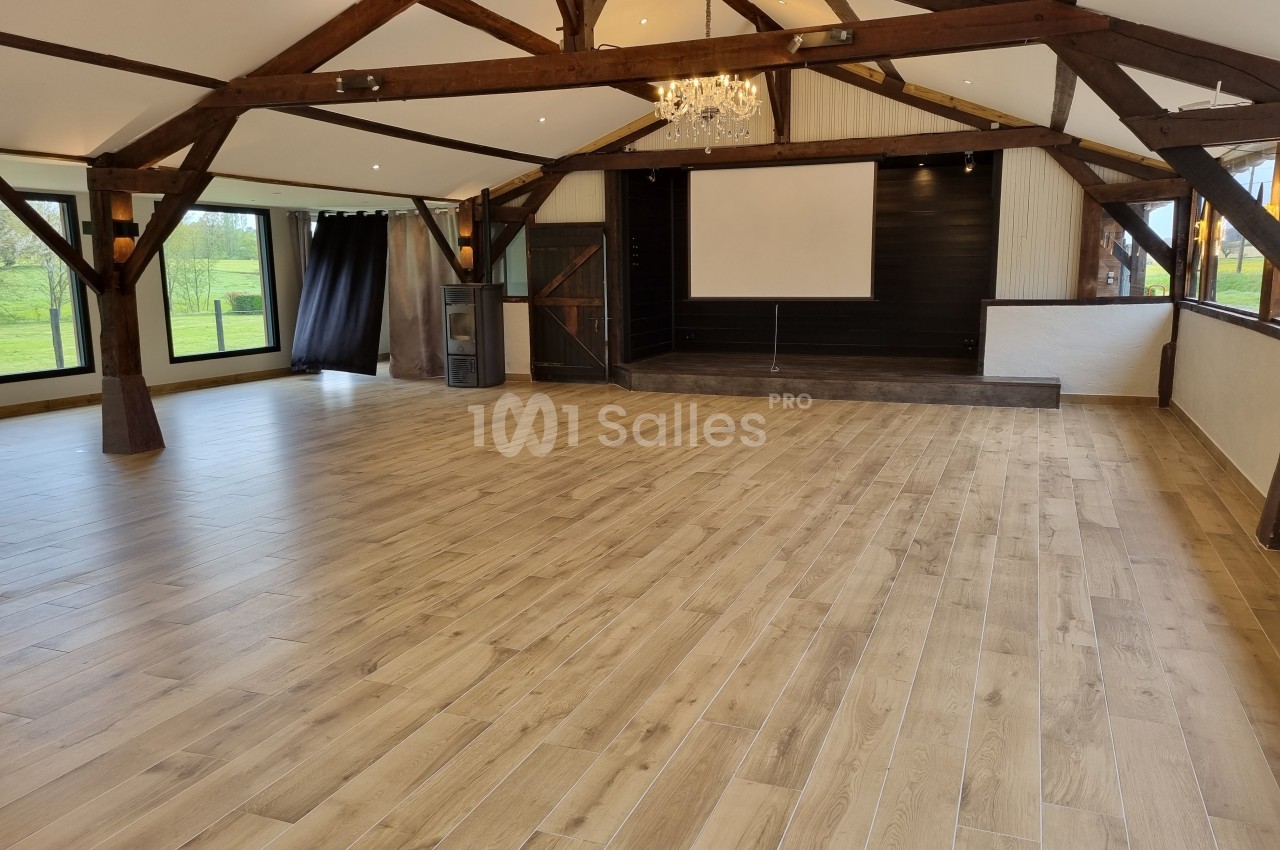Salle spacieuse avec parquet en bois, poutres apparentes, scène surélevée et grandes fenêtres donnant sur un extérieur…