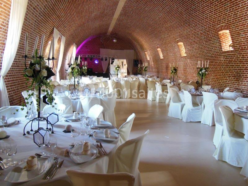 Salle voûtée en briques aménagée pour un événement, avec tables dressées, chaises blanches et décorations florales.