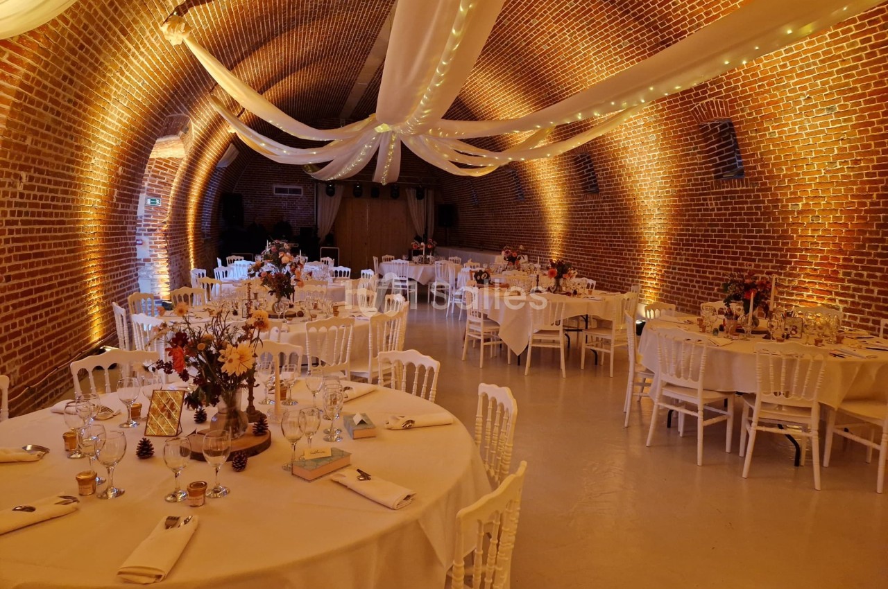 Salle voûtée en briques avec tables rondes décorées pour un événement, éclairage chaleureux et drapés suspendus.