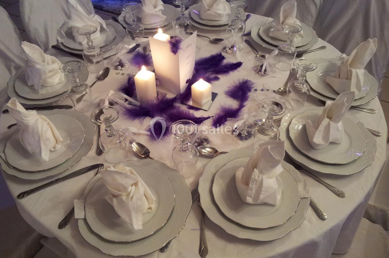 Table ronde élégamment dressée avec vaisselle blanche, serviettes pliées, bougies allumées et décorations violettes.