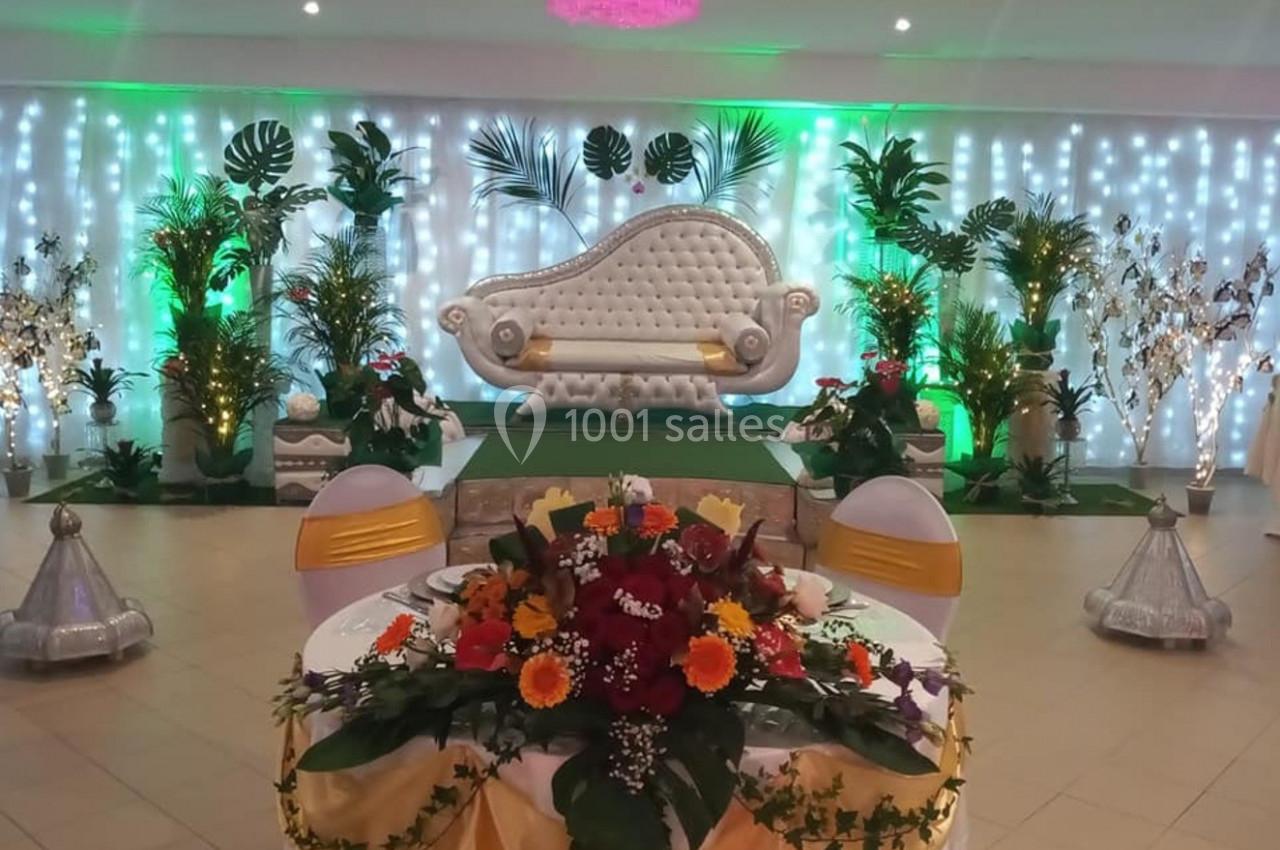 Salle décorée pour un événement avec un canapé élégant, des plantes, des lumières colorées et une table ornée de fleurs.
