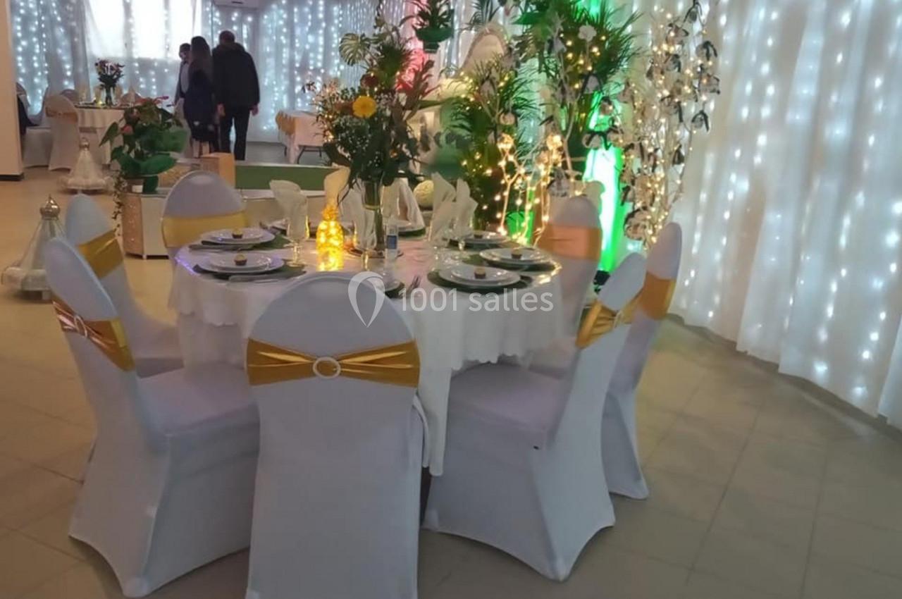 Table décorée pour un événement, entourée de chaises blanches avec nœuds dorés, éclairée par des guirlandes lumineuses.