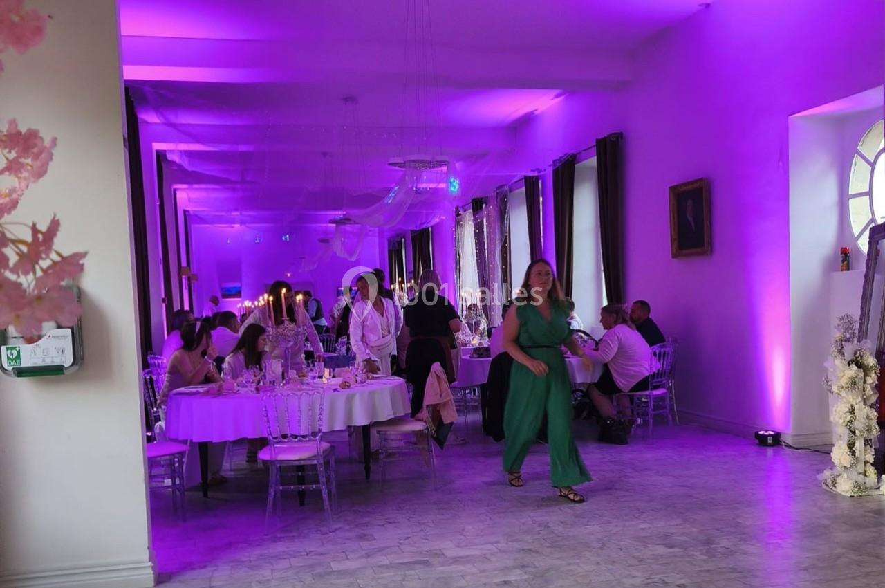 Salle de réception éclairée en violet avec des invités assis à des tables décorées et une femme en robe verte marchant.